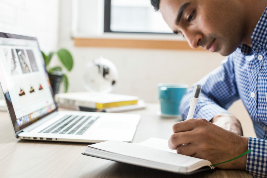 Guy writing near computer on the desk. Keep reading to learn how to Write your blog post Faster and Increase Productivity.