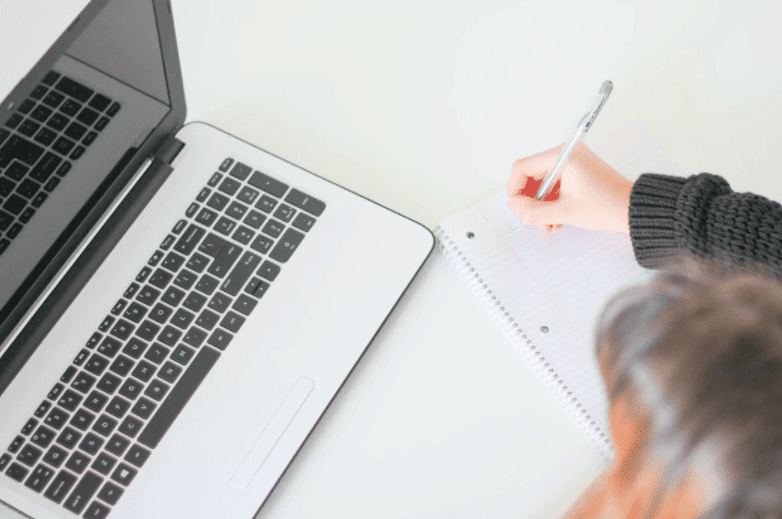 Lady making notes on white notebook for creating the best blog post workflow next to a computer.