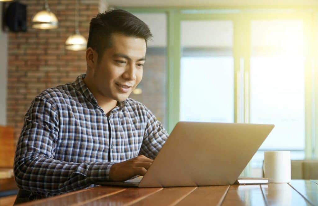 A man sitting at a table in a bright lit room with coffee and his laptop write freely in a stream of consciousness. Keep reading for tips on how to write blog posts faster.