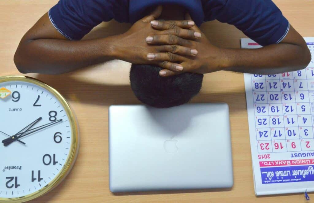 A man stressed with his head down on the table with a clock, laptop, and calendar.. Keep reading for tips on how to write blog posts faster.