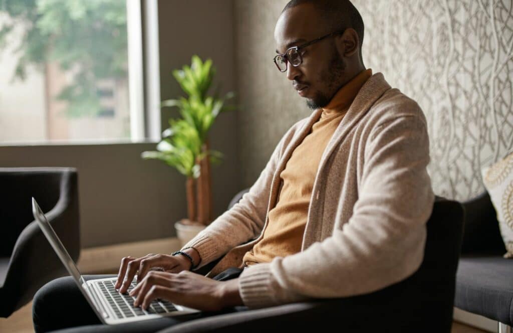 Black man sitting in a chair with laptop, making plans on the calendar to set aside for writing. Keep reading for tips on how to write blog posts faster.