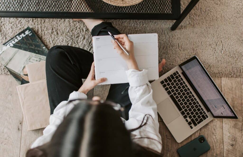 Woman sitting on the floor with journal and laptop. Keep reading to know how to write blog posts faster.