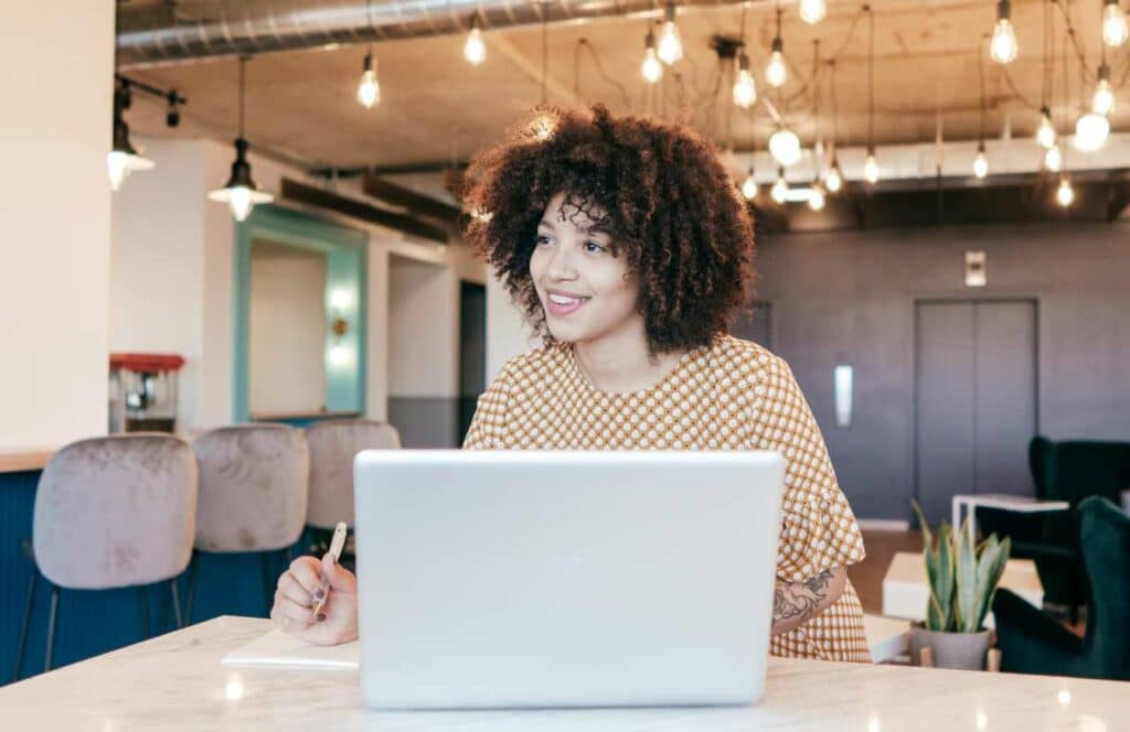 A curly haired woman sitting in a shared workspace with her laptop, pen, and paper on the desk as she brainstorms ideas of her blog brand awareness. If you want to learn how to build brand awareness for your blog, then keep reading.