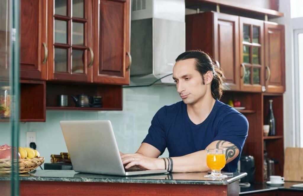 A-man-sitting-at-a-kitchen-bar-with-a-glass-of-juice-editing-a-blog-post.-Keep-reading-for-tips-on-how-to-write-blog-posts-faster.