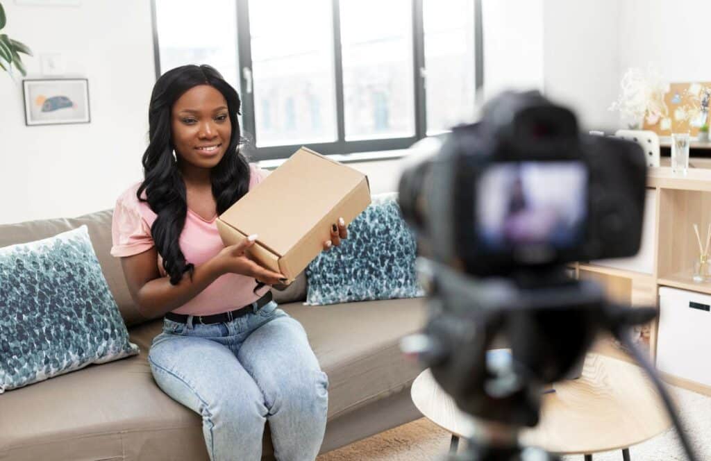 A black woman holding a box of merchandise and recording a video so she can make money blogging. Keep reading to learn how to monetize your blog from day one.