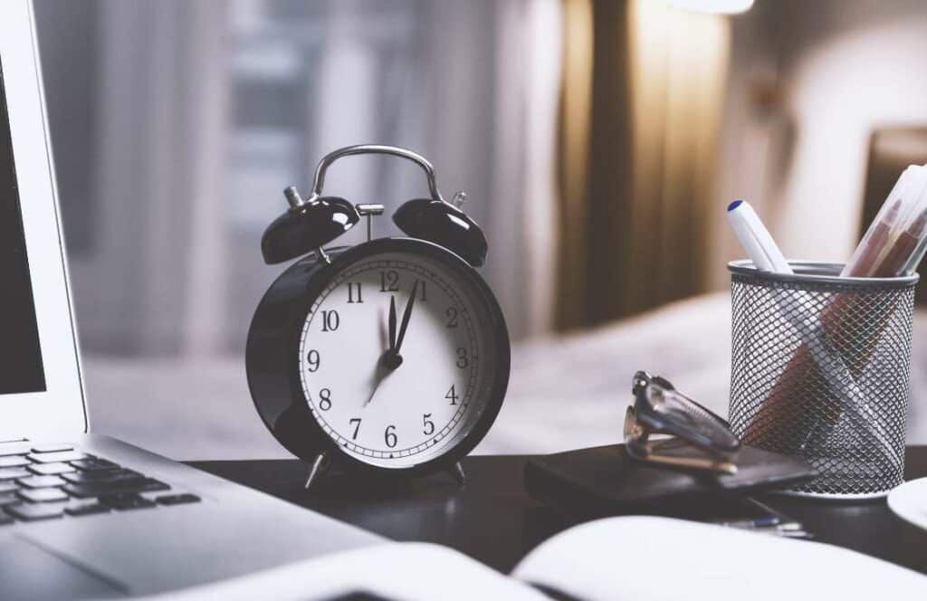 A clock, laptop, and pens on a workspace as a reminder to manage your time as a blogger. If you've ever wondered What's the point of blogging click here to read more.