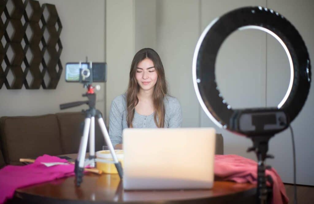 A woman sitting at a table with a camera phone, laptop, and ring light so she can create blog content. If you have ever asked "What's the point of blogging?" click here to read more."What's the point of blogging?" click here to read more.