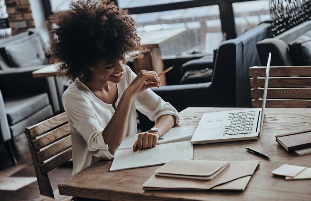 A woman sitting in a coffee shop with a notebook, pens, and a laptop. If you have ever asked "What's the point of blogging?" click here to read more.