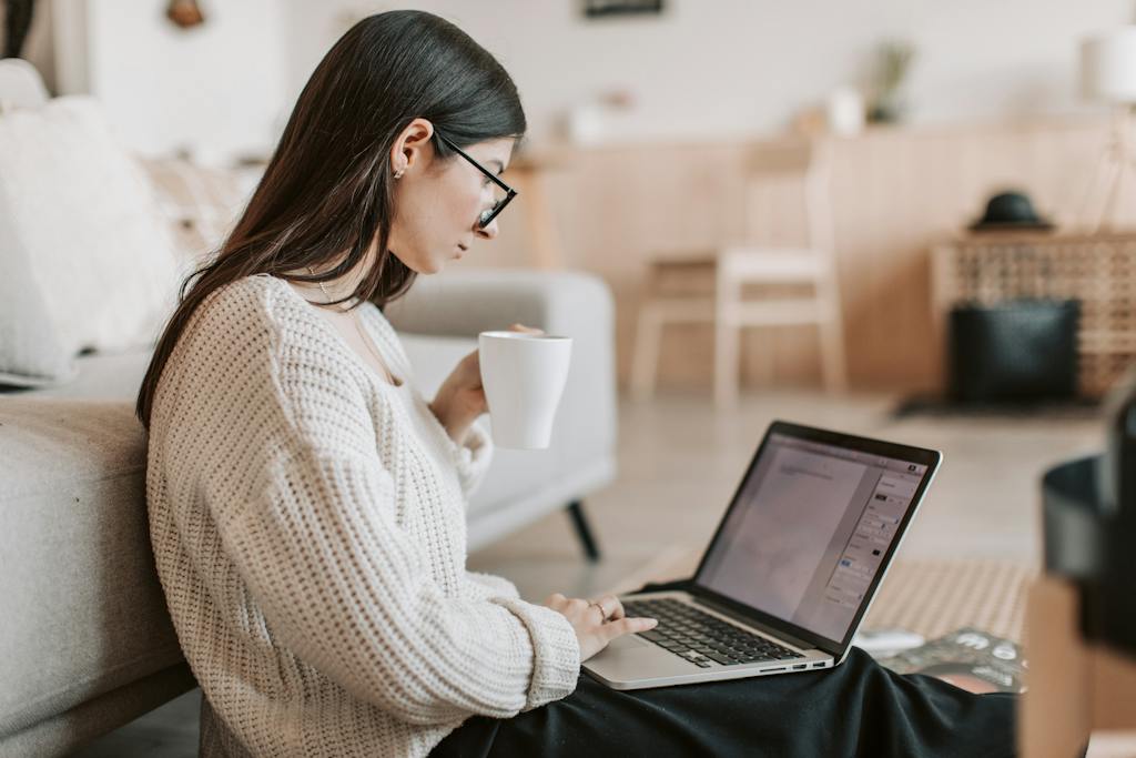 Side view of female in warm sweater and eyeglasses drinking tea from white ceramic cup while sitting on floor near sofa with netbook on legs while creating a blog post. Keep reading to learn about the best insurance affiliate marketing programs available for bloggers and content marketers. 